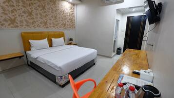 A hotel room with a bed, a wooden table with a chair, a TV mounted on the wall, and an air conditioning unit.