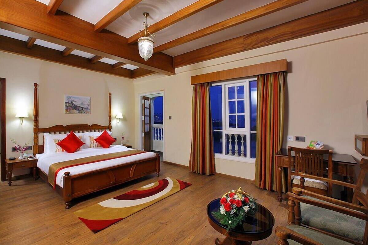 A hotel room with a large bed, wooden ceiling beams, a window with curtains, a small table with flowers, and a chair.