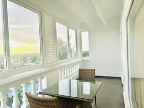 A sunlit room with a glass table and wicker chairs, large windows with shutters, and a view of greenery and buildings.