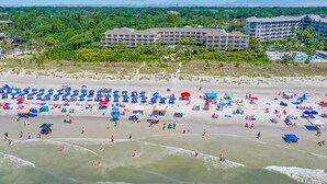 A beach with numerous umbrellas and people, a hotel in the background, and waves in the foreground. - Amazing 1-bd Beach Villa, best location Hilton Head Island -Heart of the Island! (Hilton Head Island)