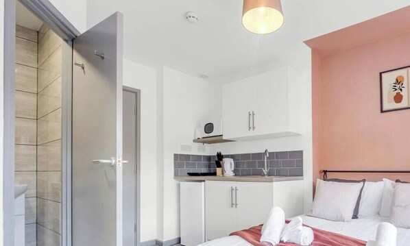 Une chambre dâhĂŽtel moderne Ă©quipĂ©e dâune douche, dâune kitchenette, dâun lit avec des serviettes et dâun luminaire au plafond.