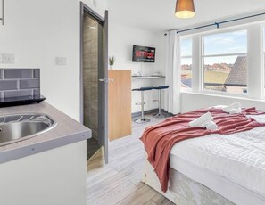 Habitación de hotel compacta con kitchenette, una cama con una colcha rosa, un televisor montado en la pared y una ventana con vistas a edificios.