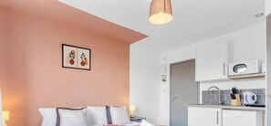 Un dormitorio con cama, almohadas, una lámpara de noche, un cuadro en la pared y una kitchenette con microondas y utensilios.