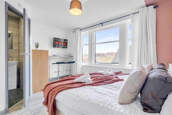 Une chambre à coucher avec un lit, une fenêtre avec des rideaux, un téléviseur fixé au mur et une vue sur l’extérieur.