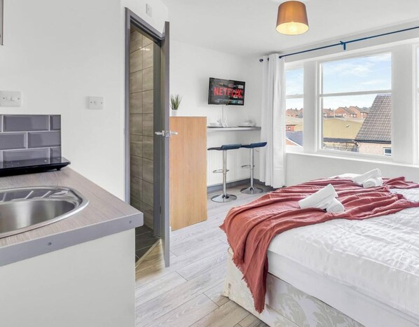 Ein kleines Hotelzimmer mit einer Küchenzeile, einem Bett mit rosa Decke, einem an der Wand befestigten Fernseher und einem Fenster mit Blick auf Gebäude.