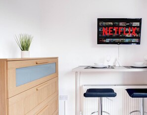 Eine moderne Küche mit einem Holzkasten, einem Blumentopf, einer Bar mit Hockern und einem an der Wand montierten Fernseher, der das Netflix-Logo zeigt.