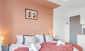 Une chambre Ă coucher comprenant un lit, des oreillers, une lampe de chevet, un tableau encadrĂ© au mur, et une kitchenette Ă©quipĂ©e dâun four Ă micro-ondes et dâustensiles de cuisine.