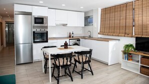 A modern kitchen with white cabinets, a stainless steel refrigerator, a microwave, and a sink. There is a dining table with black chairs and a small potted plant on the counter. - Apartamento Amplio y Tranquilo con Vistas al Mar 4 (CN)