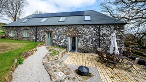 Un bâtiment en pierre doté d’un toit en pente, de panneaux solaires, d’une terrasse pavée avec des meubles d’extérieur, et d’un chemin en pierre menant à l’entrée.