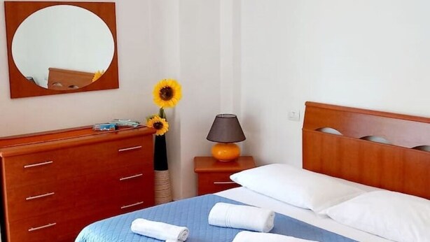 A hotel room with a bed, a wooden dresser, a mirror, a lamp, and a vase with sunflowers. - 2 Bedroom Apartment in Pisa (Pisa)