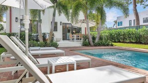 A poolside area with lounge chairs and a table, surrounded by palm trees and a building with a modern design.