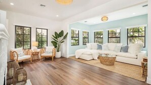 A spacious living room with a white sofa set, a coffee table, wicker chairs, and large windows offering a view of greenery.