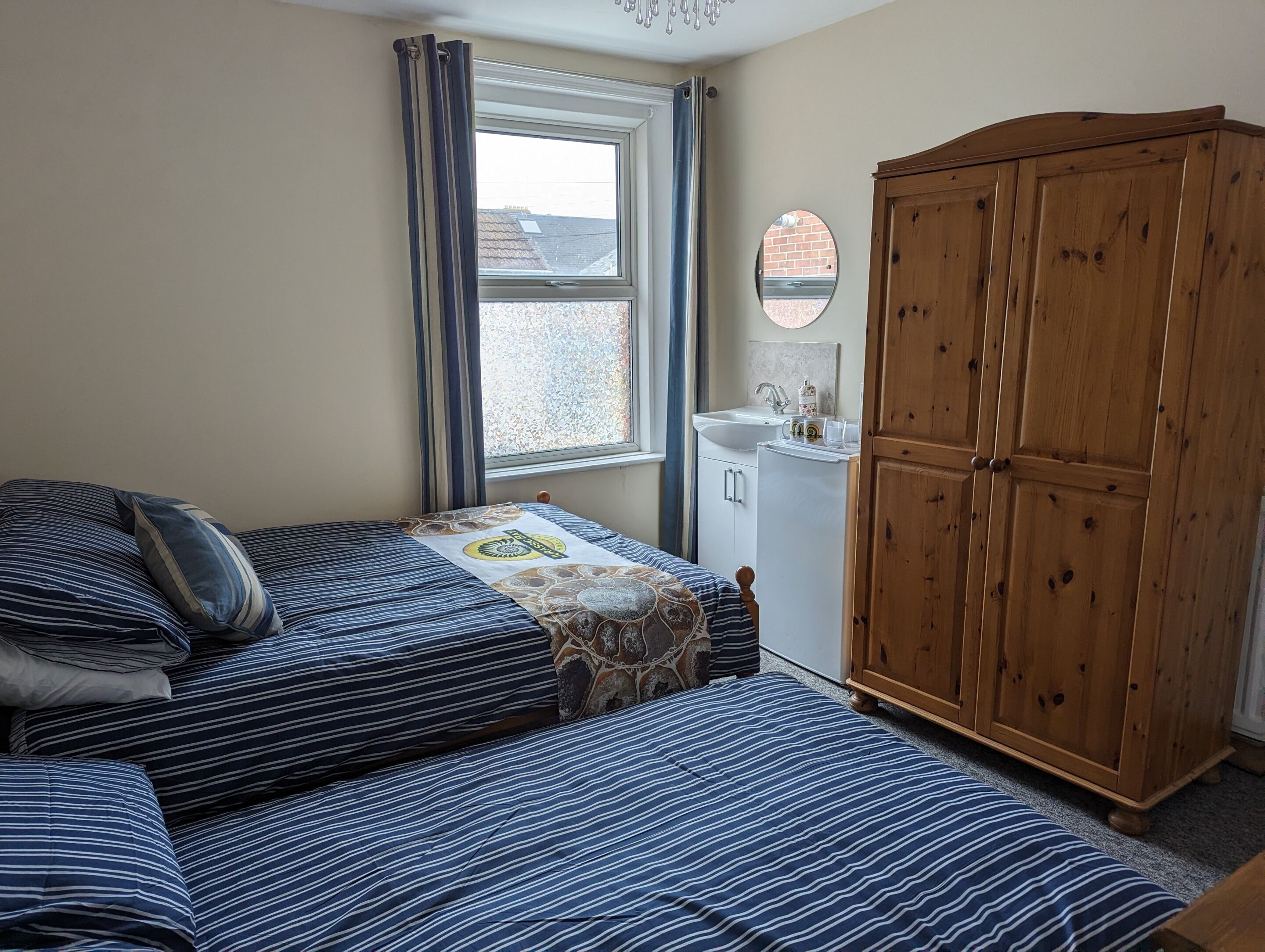 Een slaapkamer met twee bedden, een houten garderobe, een raam met gordijnen, en een kleine badkamer met een wastafel en spiegel.
