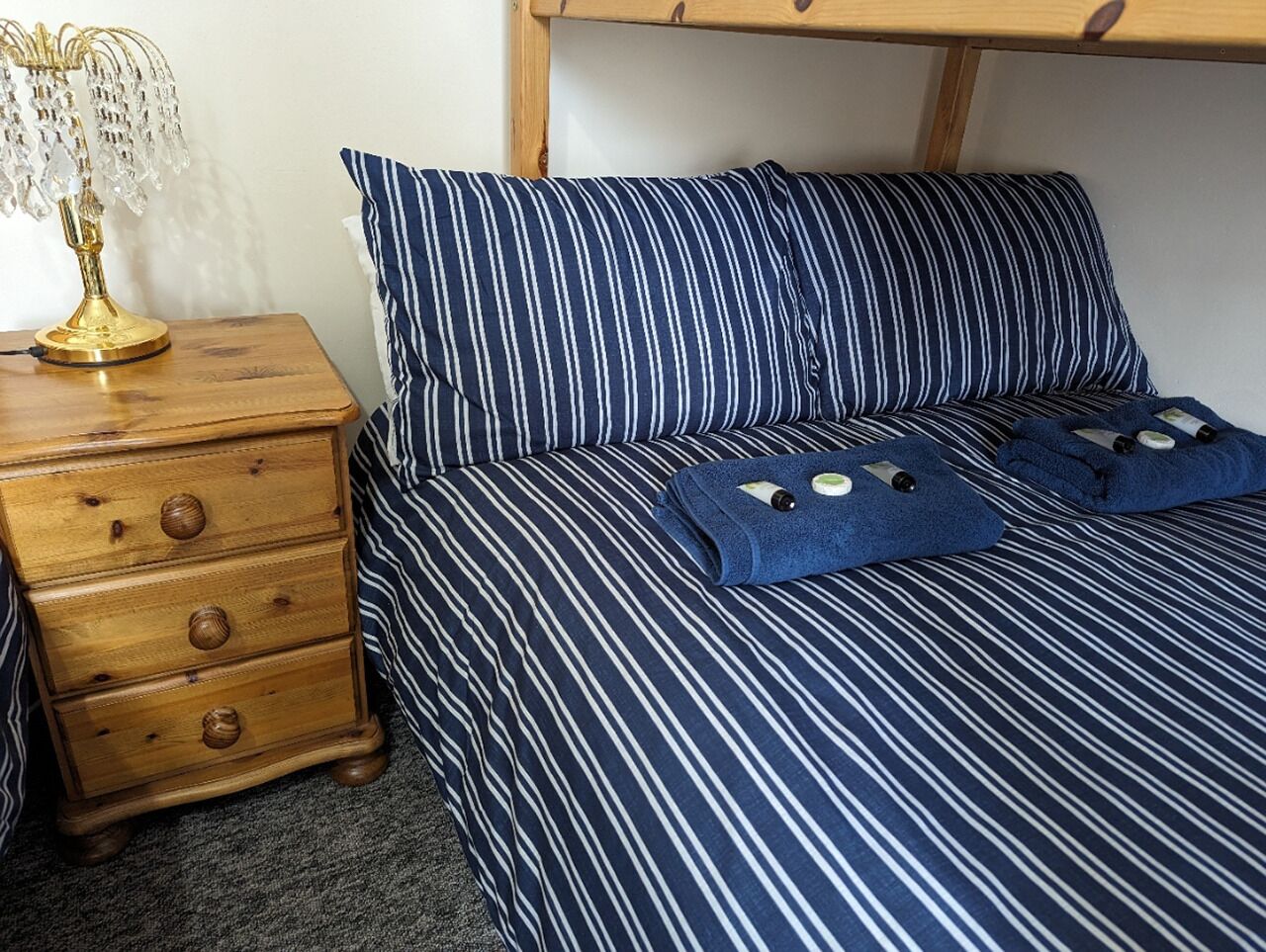 Uma cama com roupa de cama listrada e duas almofadas, um criado-mudo de madeira com gavetas e uma luminária.