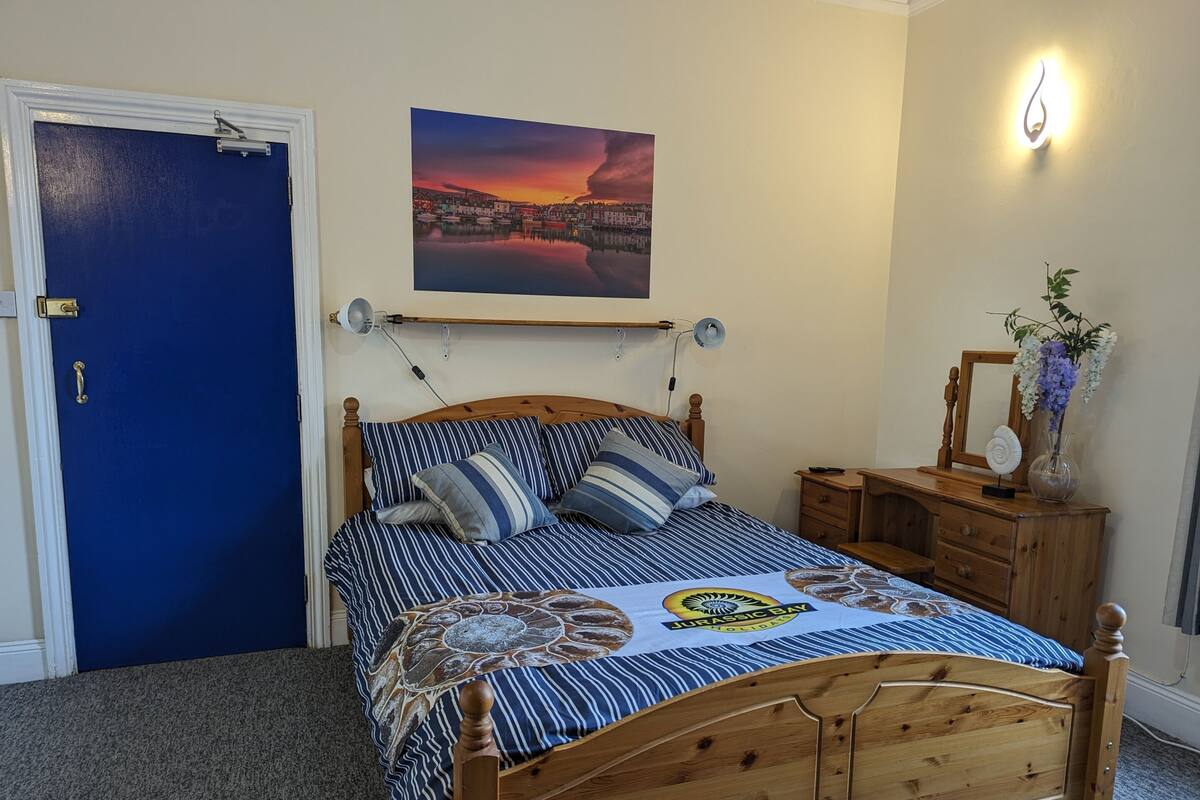 Un dormitorio con cama de madera, una puerta azul, un cuadro con una vista urbana, una mesita de noche con lámpara y un espejo.