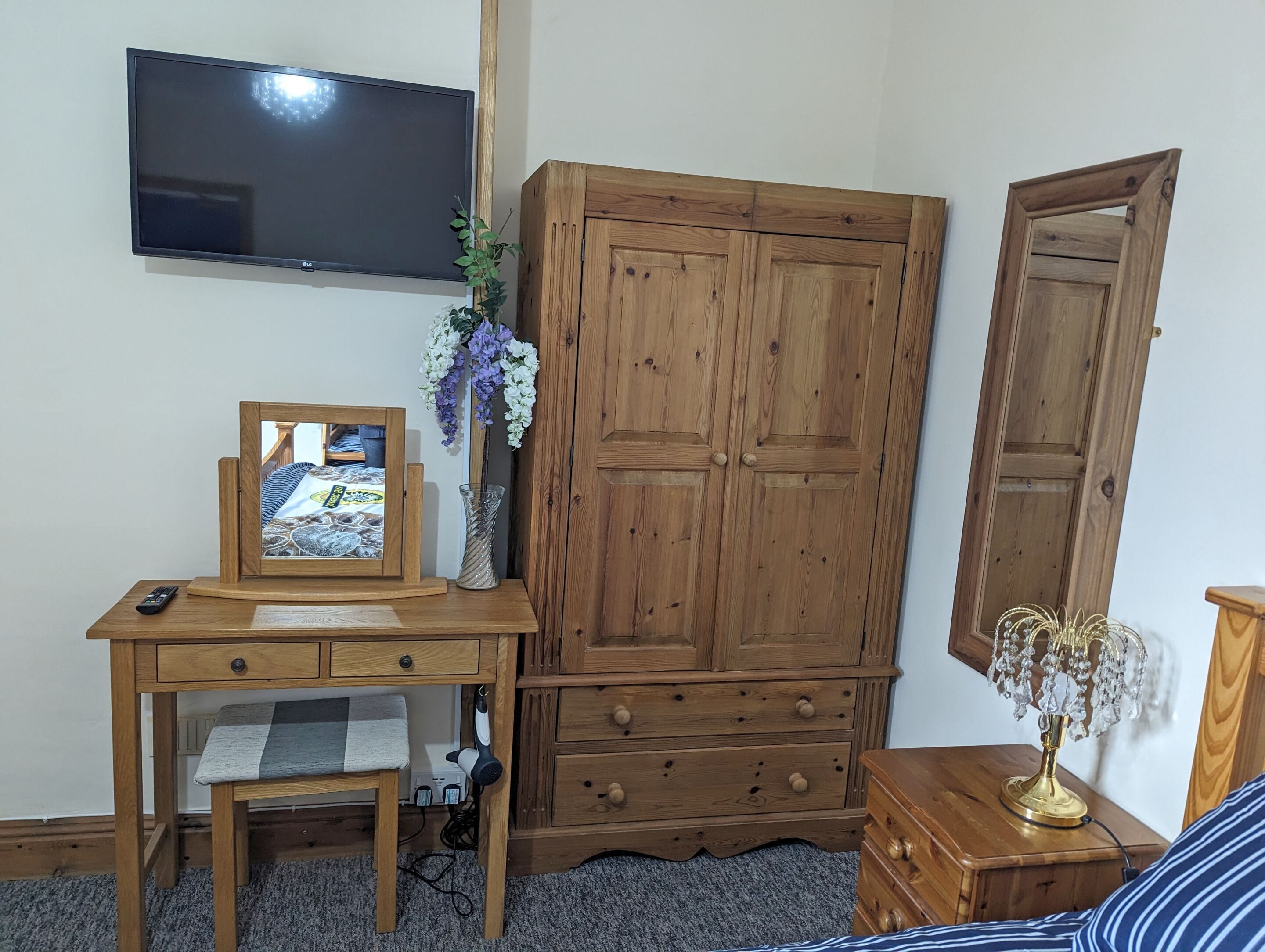 A bedroom with a wooden desk, a TV mounted on the wall, a wardrobe, a bedside table with a lamp, and a bed with striped bedding.