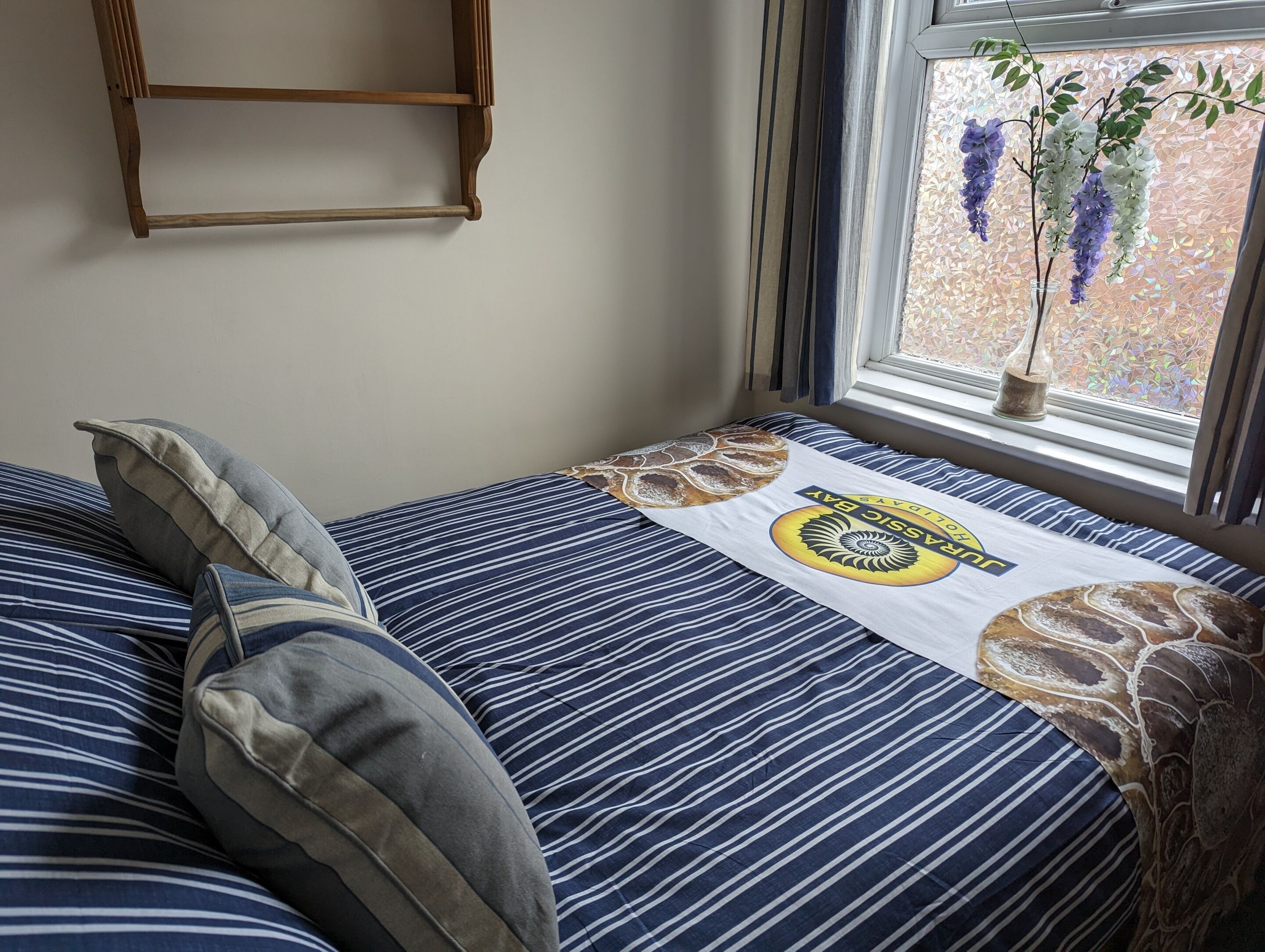 Een bed met gestreepte lakens, een kussen, een decoratieve handdoek en een raam met uitzicht op bloemen en een bakstenen muur.