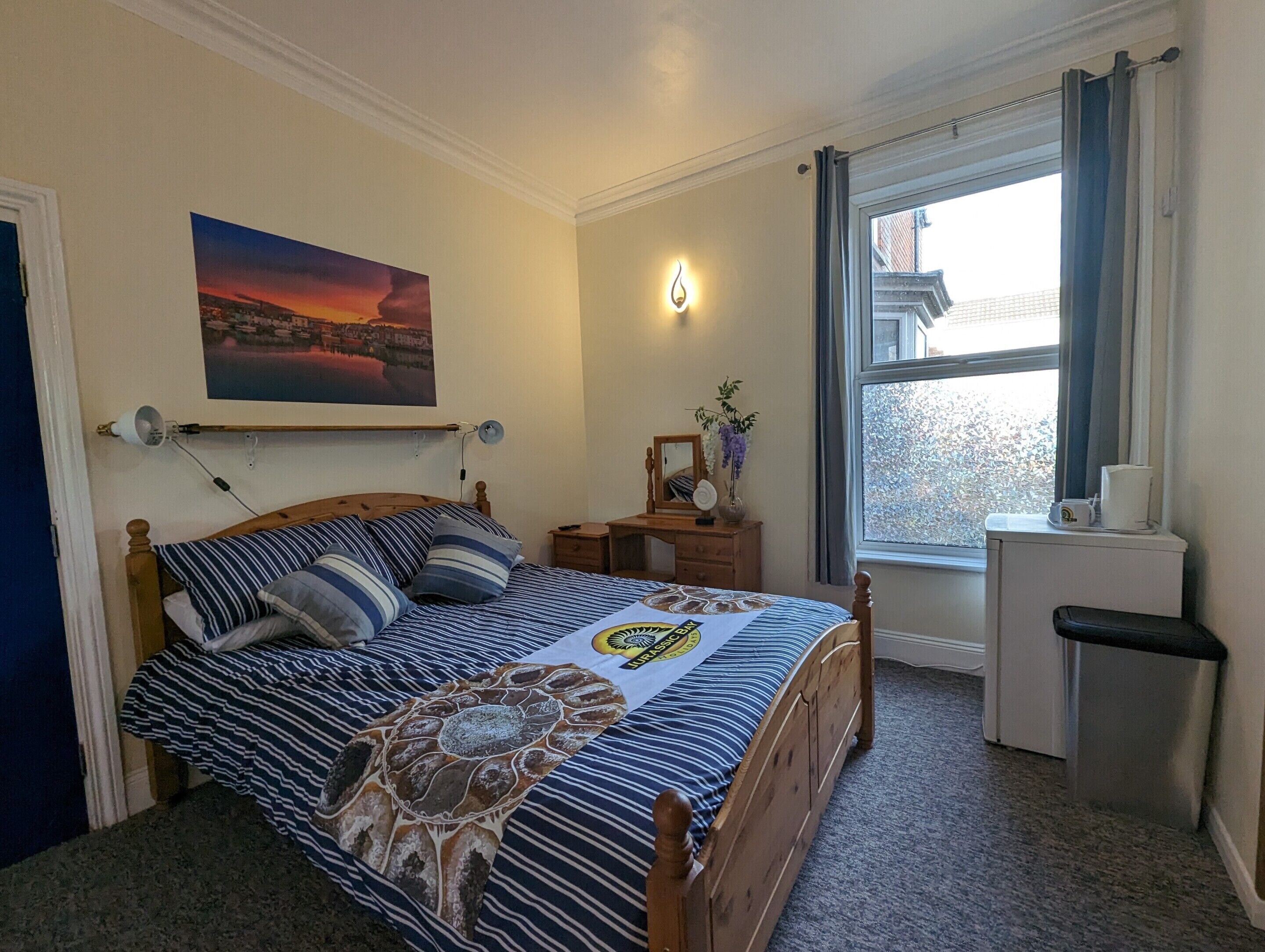 A bedroom with a bed, a bedside table, a dresser, a window with curtains, and a painting on the wall.