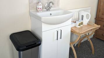 Un baño con un lavamanos blanco, un espejo, un cubo de basura y una mesita de madera con una tetera y tazas.