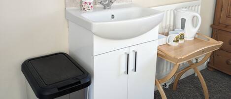 A bathroom with a white sink, a mirror, a trash bin, and a wooden table with a kettle and cups.