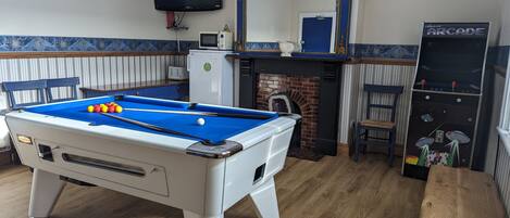 A pool table with balls, a television, a mirror, a fireplace, and a jukebox in a room with wooden flooring and patterned walls.
