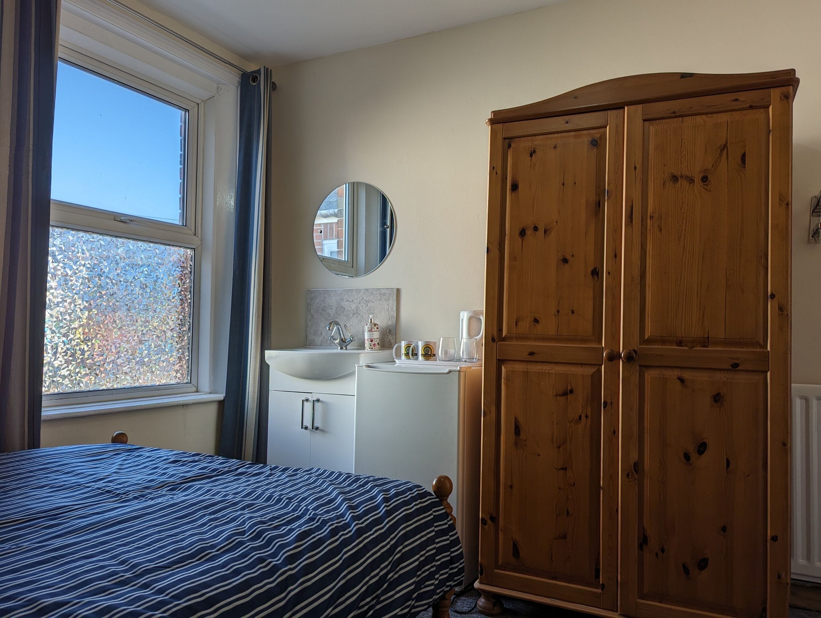 A bedroom with a bed, a wooden wardrobe, a white dresser, a mirror, and a window with curtains.