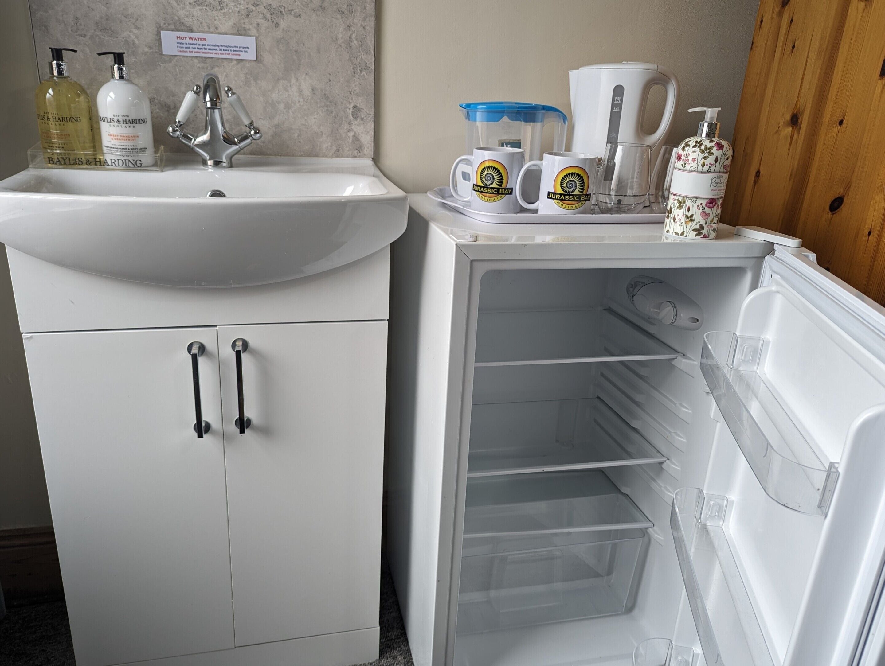 Un baño con lavamanos, dos botellas de jabón para manos, cafetera y refrigerador con cajones transparentes.
