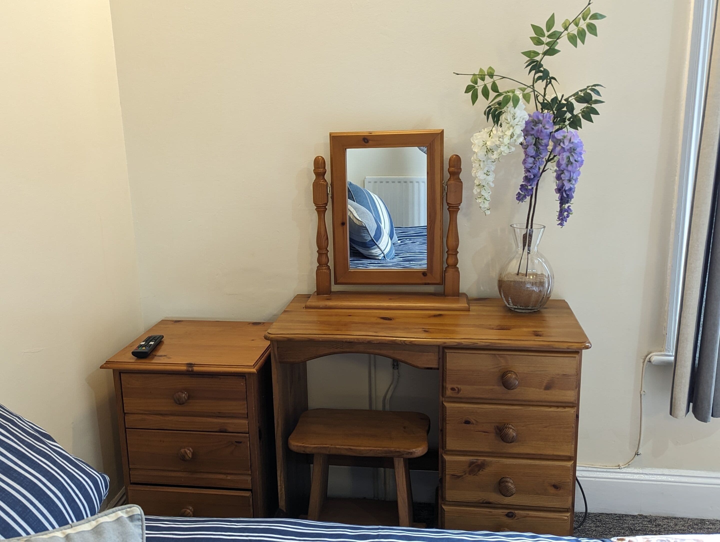 Un bureau en bois avec un miroir, un vase rempli de fleurs et un lit avec une literie à rayures.