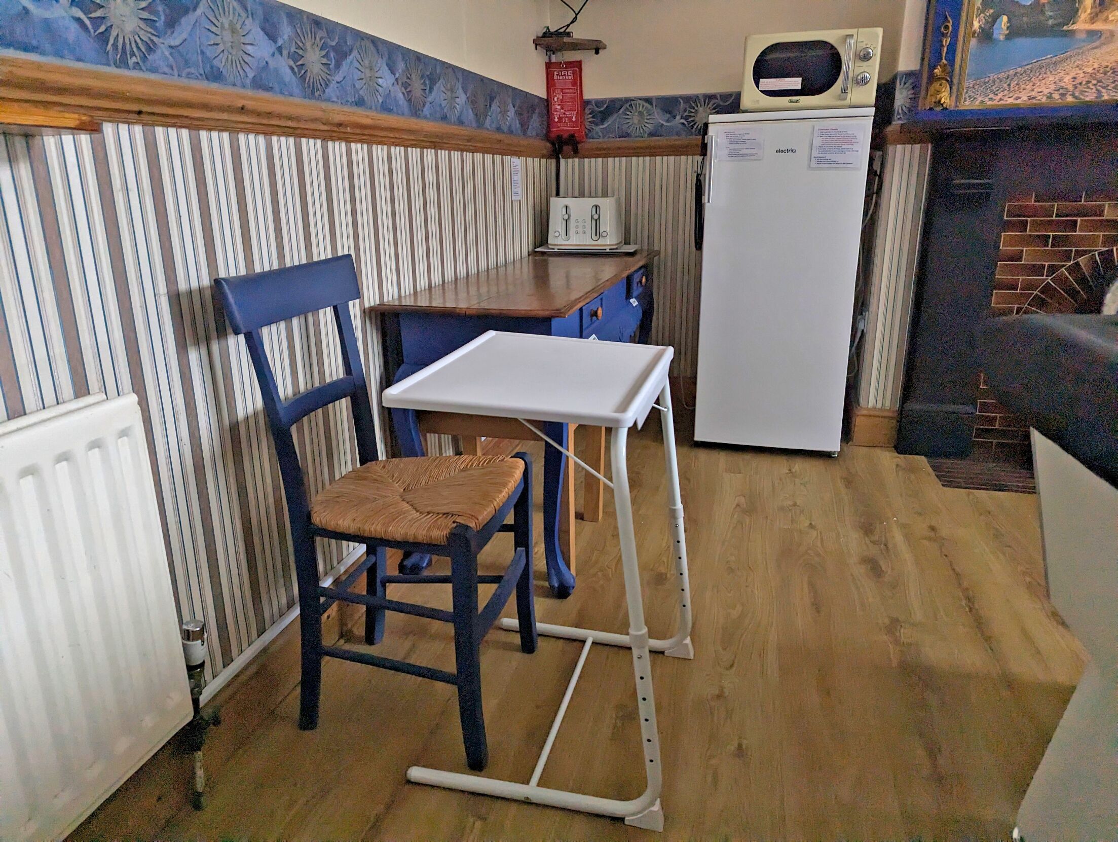 A compact kitchen with a striped wall, a wooden table, a white refrigerator, and a blue chair.