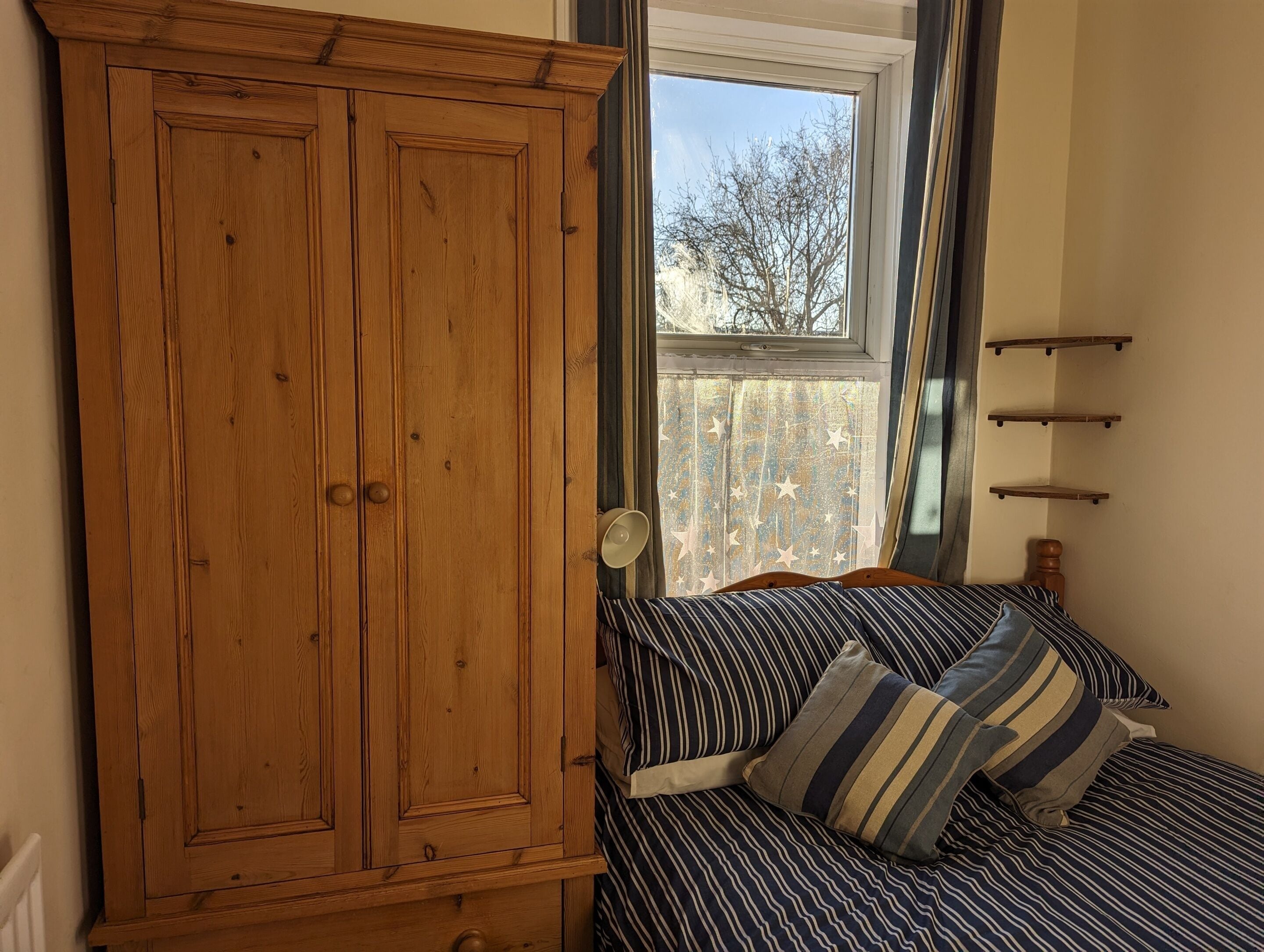 A bedroom with a wooden wardrobe, a bed with striped bedding, and a window with a view of trees.