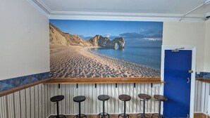 A bar area with a wooden counter and five stools, a blue door, and a mural of a beach scene. - Jurassic Bay Holidays (Weymouth)