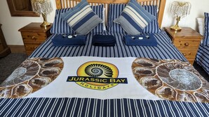 A bed with striped bedding, two pillows, and a blanket with a Jurassic Bay logo. - Jurassic Bay Holidays (Weymouth)