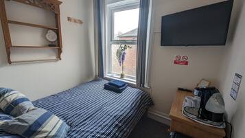 Una habitación pequeña y ordenada con una cama, un escritorio con cafetera, un televisor y una ventana que ofrece vistas a edificios.