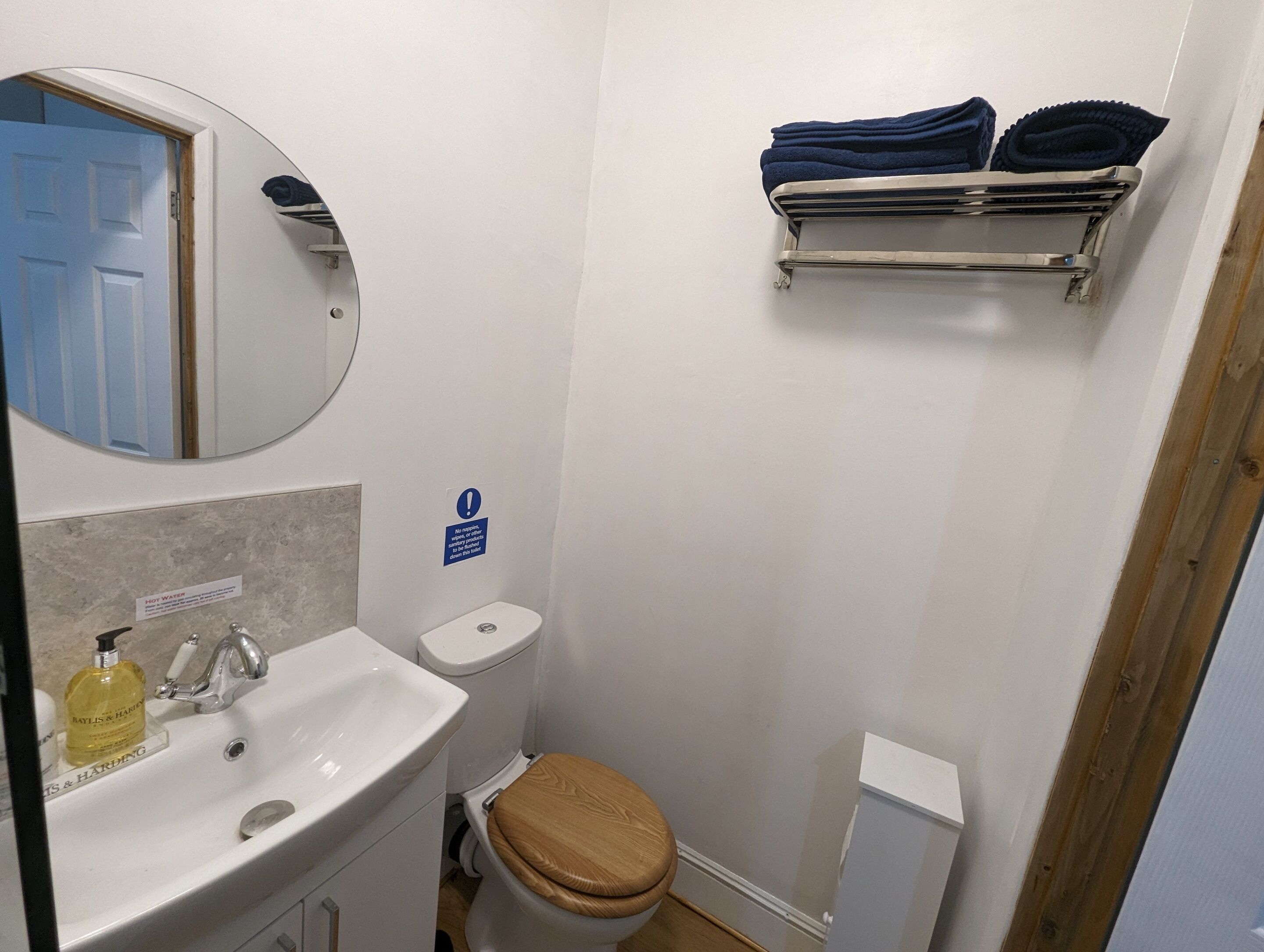 A small bathroom with a sink, toilet, and mirror. There is a sign indicating no smoking, and towels are hung on a rack.