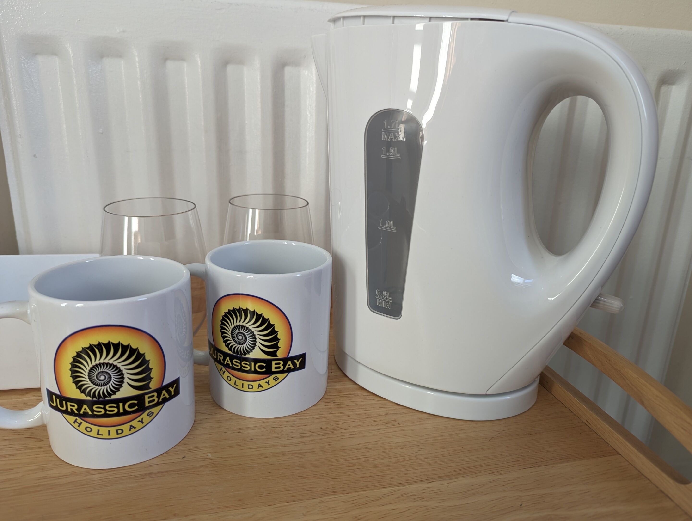 A white kettle, two mugs with a dinosaur design, and two clear glasses on a wooden surface.