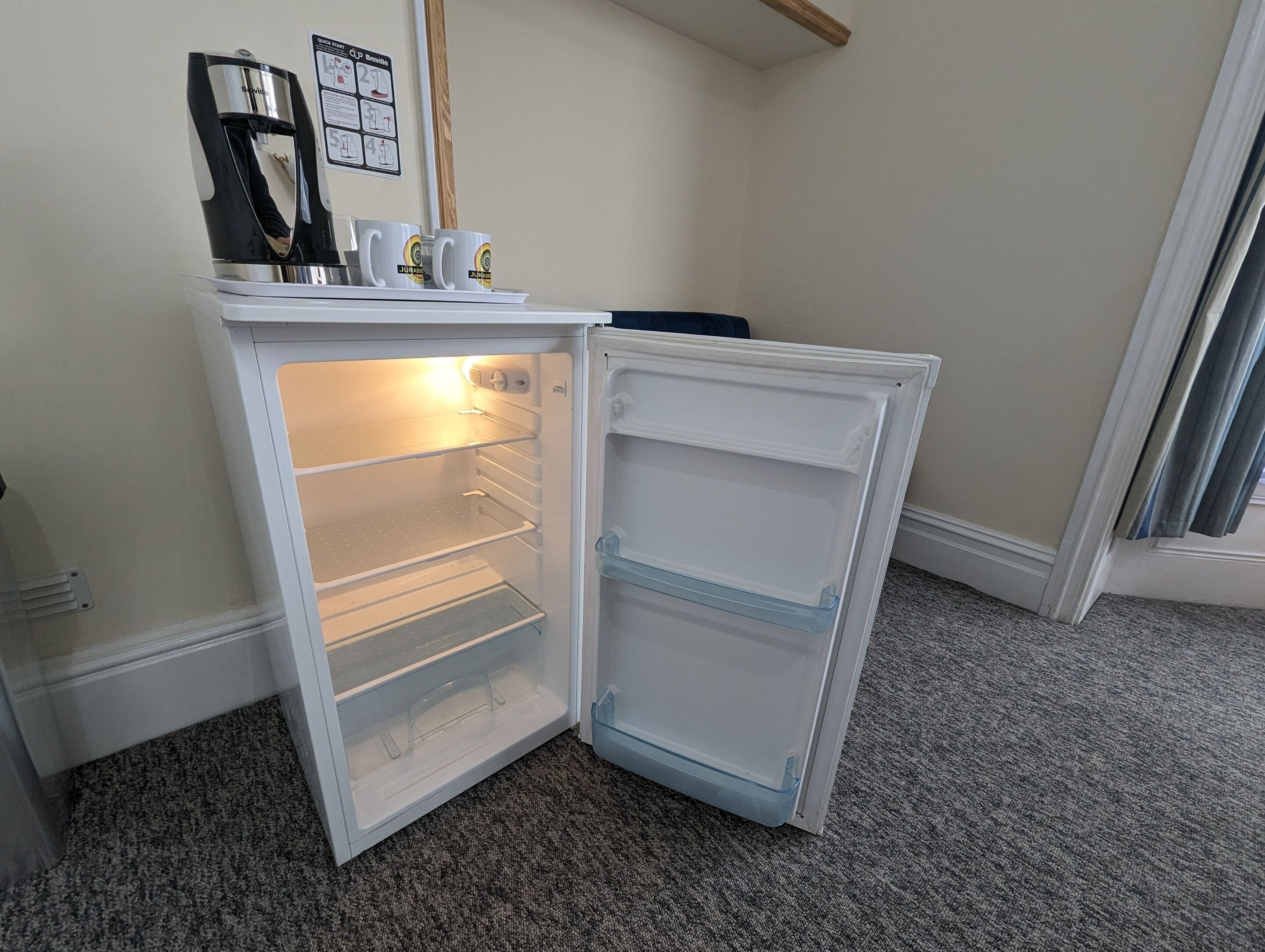 Un refrigerador compacto con dos estantes y un compartimento congelador, ubicado en un rincón de una habitación con piso alfombrado y techo con vigas de madera.
