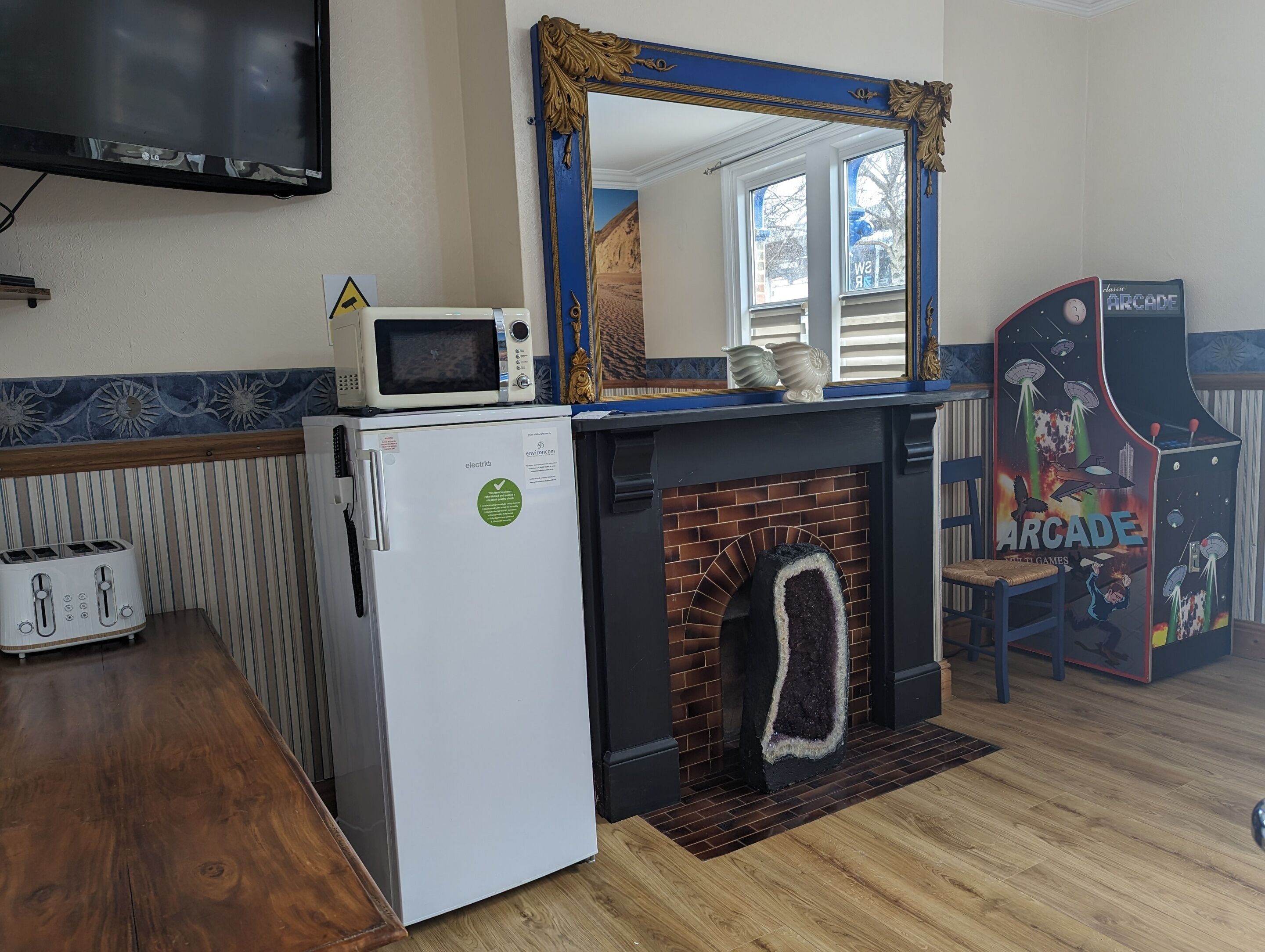 A compact room with a microwave on a small fridge, a toaster, a fireplace with a rug, and a retro arcade machine.