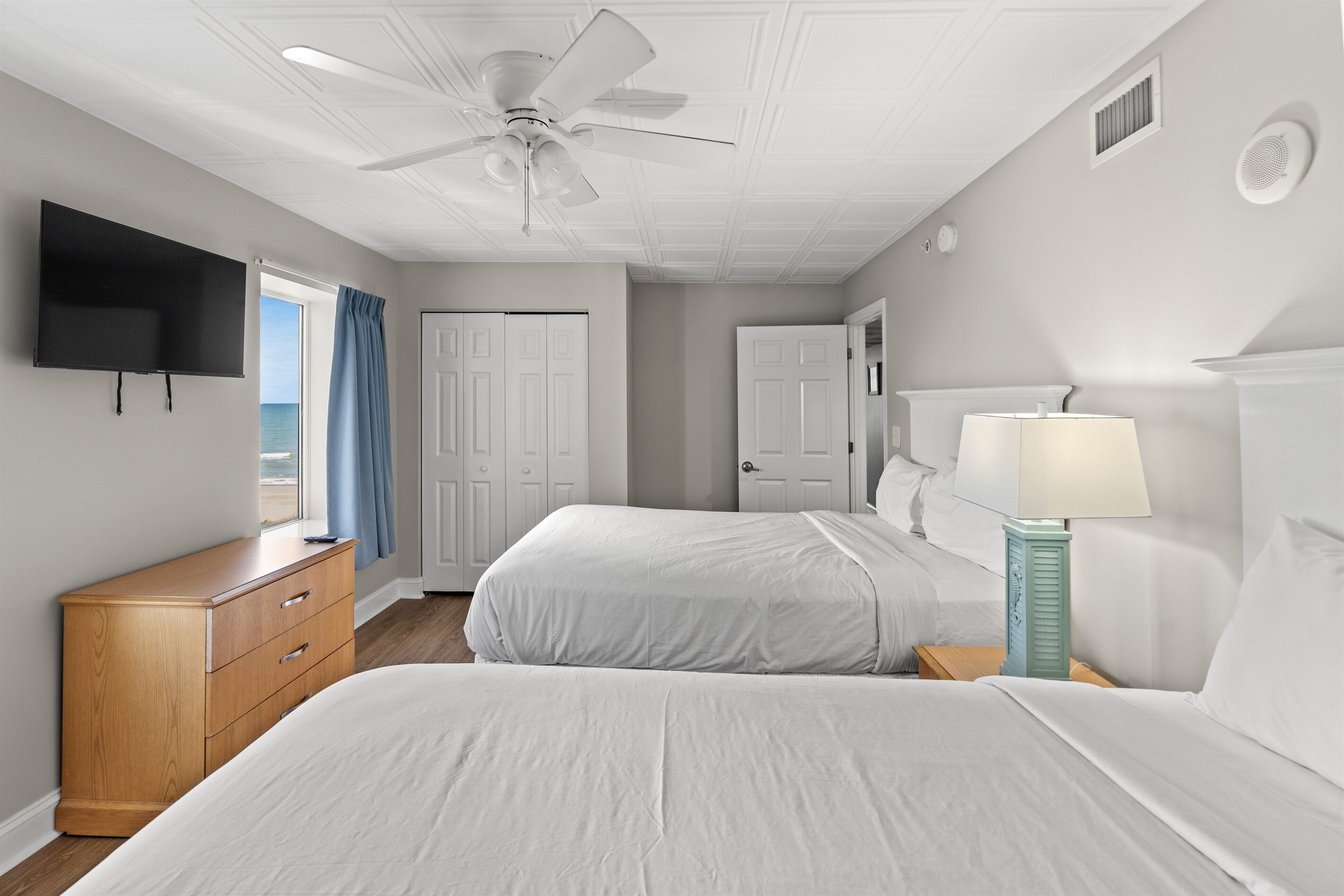 Une chambre d’hôtel avec deux lits, un ventilateur de plafond, une télévision, une commode en bois et une lampe.