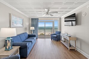 Sala de estar con un sofá azul, una mesita con una lámpara, una silla, un televisor y vistas a la playa a través de un ventanal.