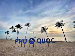 A beach with palm trees and a sailboat in the distance. - Palmera Villa Phu Quoc (Phu Quoc)