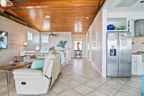 A spacious living area with a beige sofa, a dining table, and a kitchen area with a refrigerator. - “Beach Views—Shell Yeah! Beach House is your stylish, fun-filled coastal escape with private bathrooms & neighborhood pool-Just steps from the sand!” (Port Aransas)