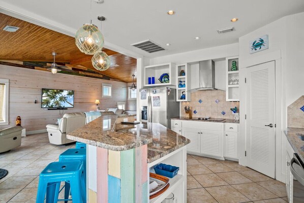 A modern kitchen with white cabinetry, a granite countertop island, and a dining area with a white sofa and blue stools. - “Beach Views—Shell Yeah! Beach House is your stylish, fun-filled coastal escape with private bathrooms & neighborhood pool-Just steps from the sand!” (Port Aransas)