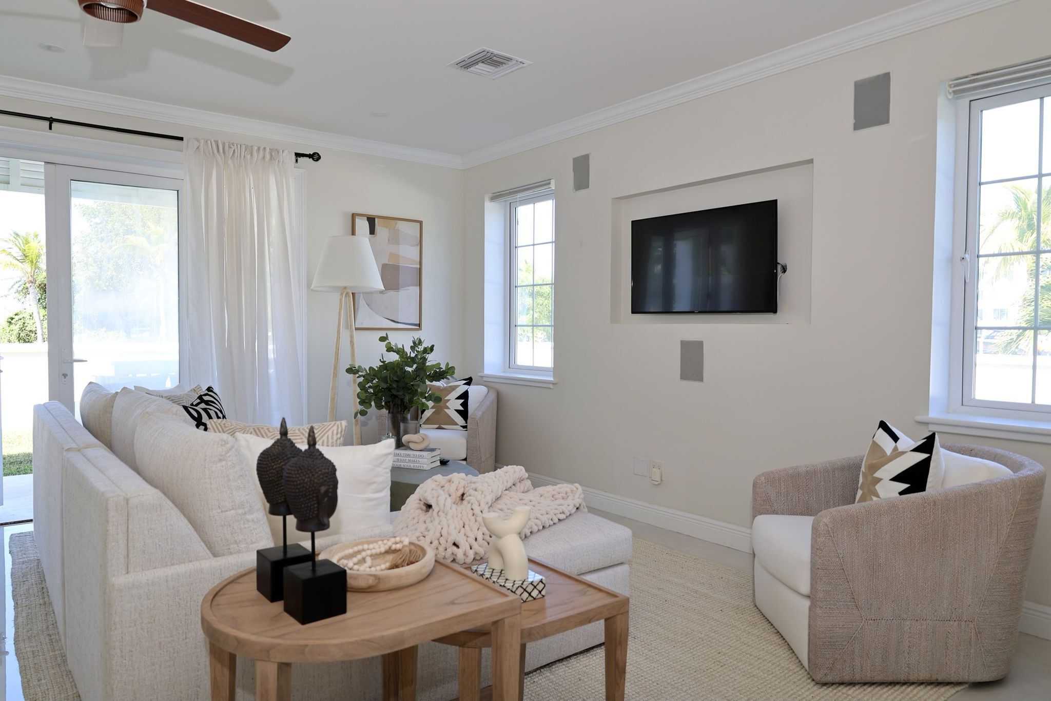 A living room with a sofa, armchair, and coffee table, a flat-screen TV mounted on the wall, and large windows with curtains.