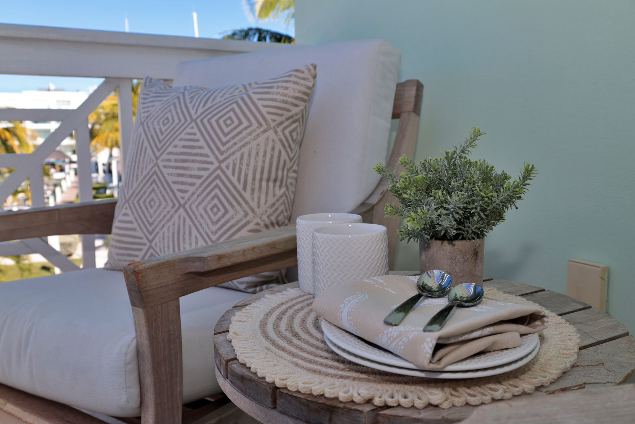 A wooden chair with a cushion and a patterned pillow, a small table with a plant, a rolled-up towel, and a plate with cutlery.