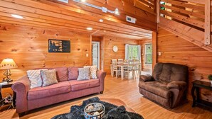 A cozy cabin interior with a wooden ceiling, a sofa, a chair, a dining table, and a fireplace.
