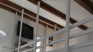 A loft-style bedroom with a wooden ceiling, a TV mounted on the wall, a ladder, and a window.