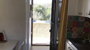 A narrow kitchen with a tiled floor, a white table with chairs, and a view of the outside through an open door.
