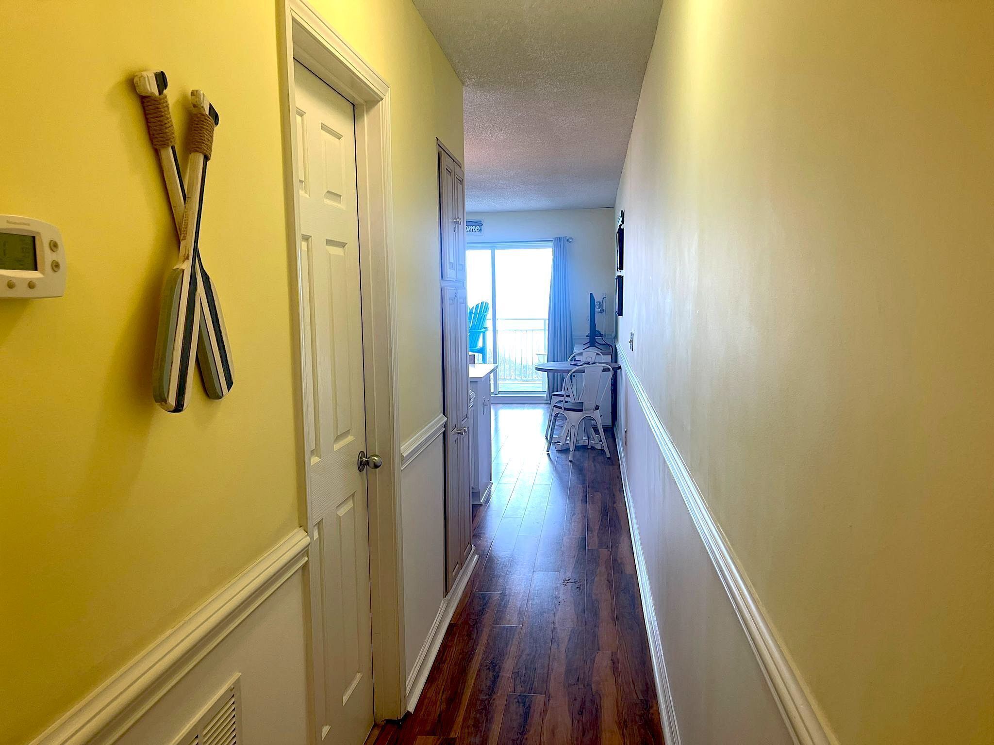 A narrow hallway with wooden flooring, a white door, and a view into a room with a balcony.