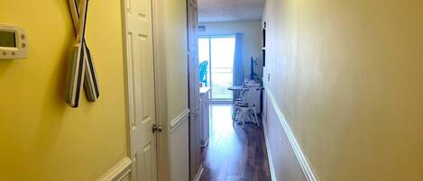 A narrow hallway with wooden flooring, a white door, and a view into a room with a balcony.
