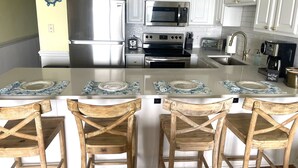 A kitchen with white cabinets, a stainless steel refrigerator, a microwave, an oven, and a coffee maker. There are four wooden chairs with a cross-back design. - Oceanfront ! All linens included ! Stunning view ! 2BR/2BA.  WindJammer Condos (Ocean Isle Beach)